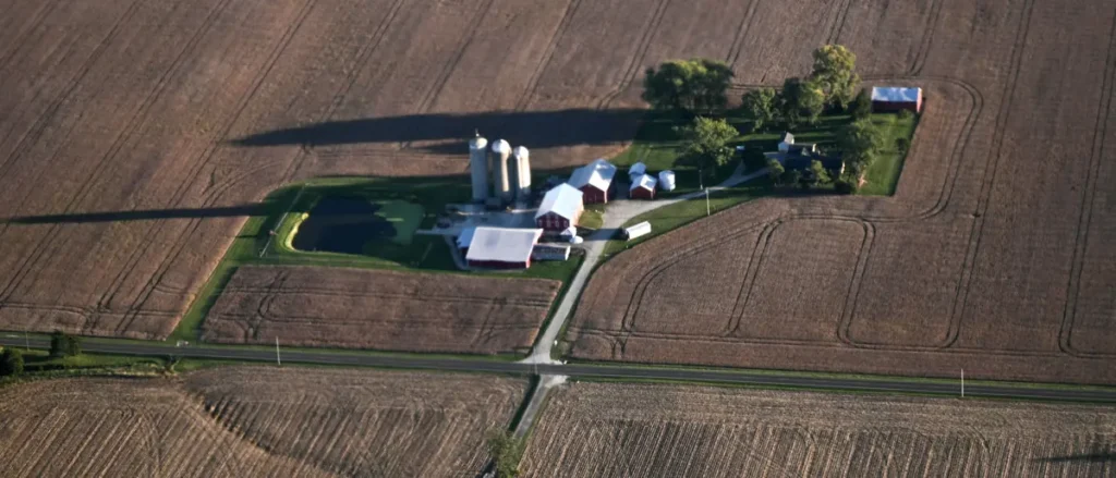 The image shows an aerial view of a rural farm with silos, barns, and a pond surrounded by plowed fields and roads.