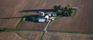 The image shows an aerial view of a rural farm with silos, barns, and a pond surrounded by plowed fields and roads.