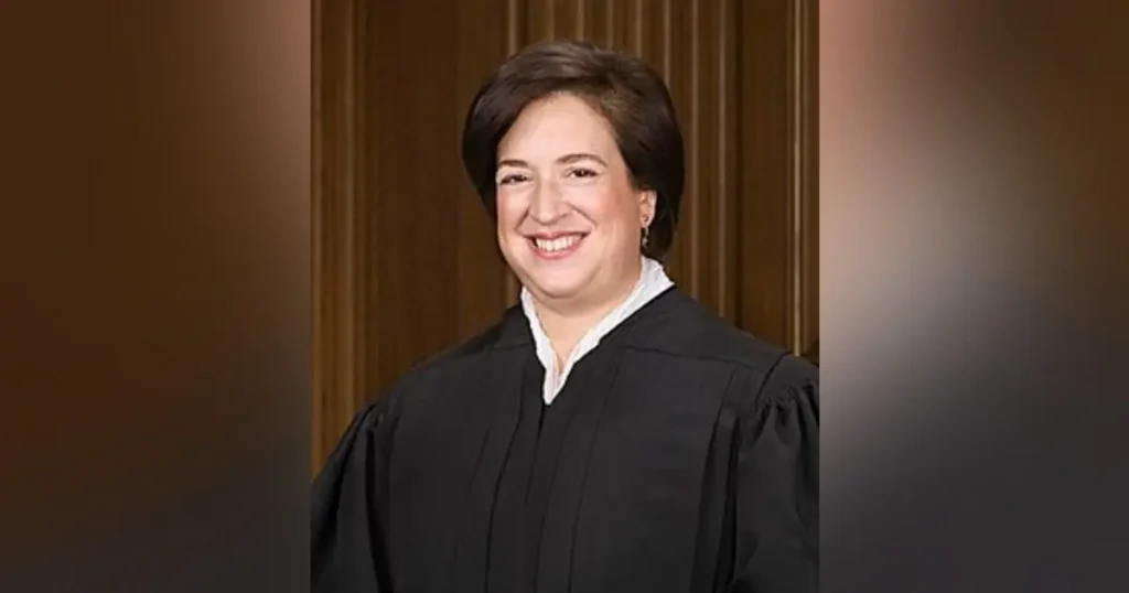The image shows a person in a black judicial robe, standing in front of a wooden panel background.