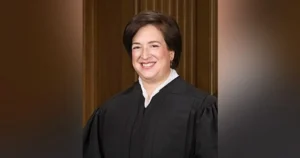The image shows a person in a black judicial robe, standing in front of a wooden panel background.