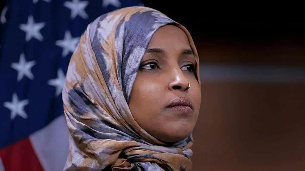 The image shows a person wearing a patterned hijab, with an American flag in the background.