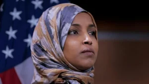 The image shows a person wearing a patterned hijab, with an American flag in the background.