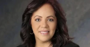 The image shows a woman with dark, wavy hair, wearing a black top and hoop earrings, against a neutral background.