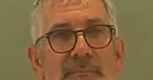 The image shows a close-up of a person wearing glasses and a mustache, with a neutral expression.