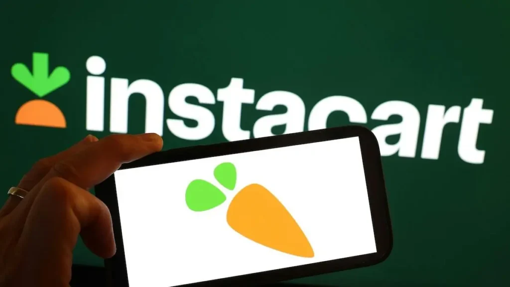 The image shows a hand holding a smartphone displaying the Instacart logo, with the Instacart brand name in the background.