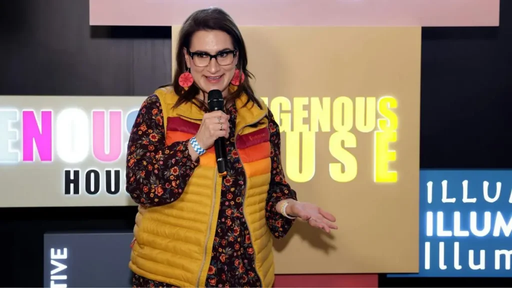 A woman is speaking into a microphone on stage, with a colorful backdrop featuring the words "INDIGENOUS HOUSE" and "ILLUMINATE."