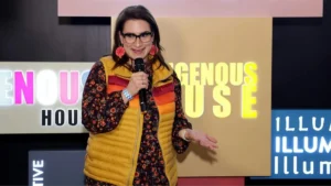 A woman is speaking into a microphone on stage, with a colorful backdrop featuring the words "INDIGENOUS HOUSE" and "ILLUMINATE."