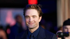 The image shows a person dressed in a formal black suit and tie, smiling at the camera in what appears to be a public or event setting.