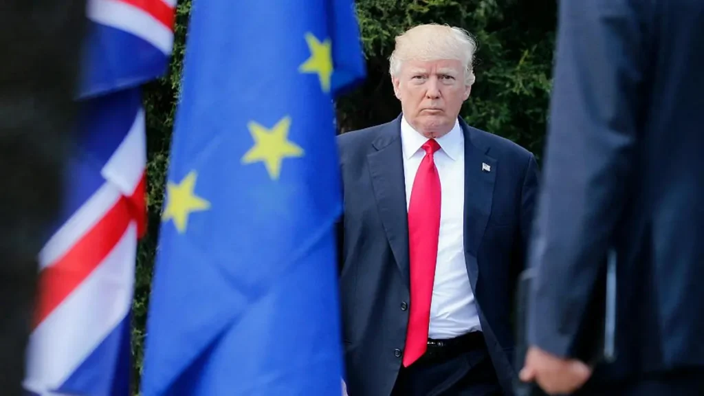 The image shows a man in a suit and red tie standing in front of European Union and British flags.