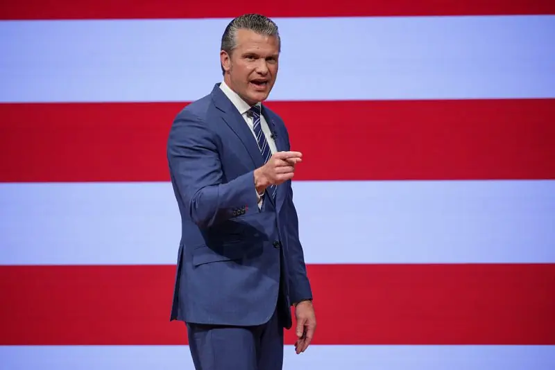 A man in a suit gestures while speaking in front of a backdrop featuring red and white stripes.