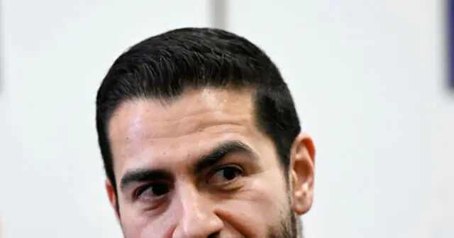 The image shows a close-up of a person with short dark hair, wearing a suit, and looking to the side with a serious expression.
