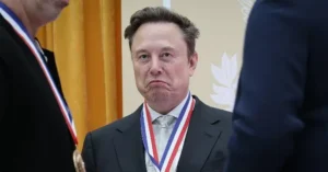 The image shows a man in a suit and tie, wearing a medal with a red, white, and blue ribbon, standing in a formal setting.