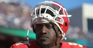 The image shows a football player wearing a red helmet and jersey, standing on a field with a blurred stadium in the background.