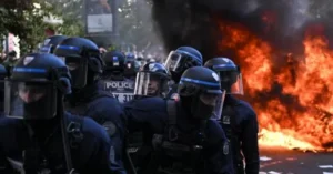 A group of police officers in riot gear stand in front of a large fire during a protest.