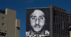 A large billboard featuring a black-and-white portrait of Colin Kaepernick with the Nike slogan "Just do it" and the phrase "believe in something, even if it means sacrificing everything" is displayed against a backdrop of urban buildings.