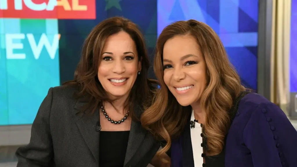 Two women are smiling and posing together on a news set, with a colorful background and the word "NEW" visible.