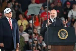 The image shows a public event where a man in a suit is speaking at a podium with the presidential seal, while another man in a suit and a "USA" hat stands beside him.