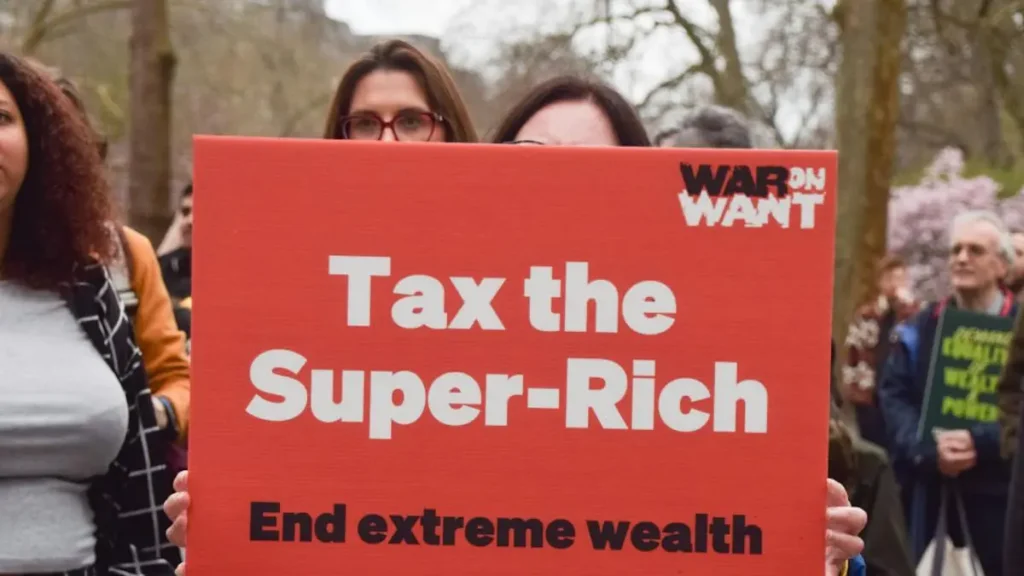 The photo shows a protestor holding a sign that reads "Tax the Super-Rich" and "End extreme wealth," advocating for higher taxes on the wealthy.