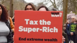 The photo shows a protestor holding a sign that reads "Tax the Super-Rich" and "End extreme wealth," advocating for higher taxes on the wealthy.