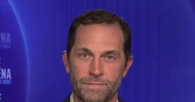 The image shows a man with a beard and short hair, wearing a suit, against a blue background with text partially visible.