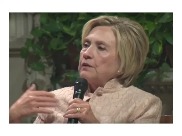 The image shows a person speaking into a microphone, with a background of greenery and a light-colored wall.