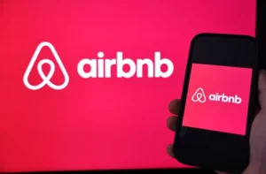 A person holds a smartphone displaying the Airbnb logo in front of a larger Airbnb logo on a screen.