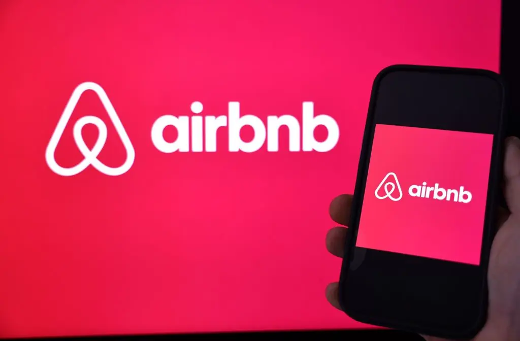 A person holds a smartphone displaying the Airbnb logo in front of a larger Airbnb logo on a screen.