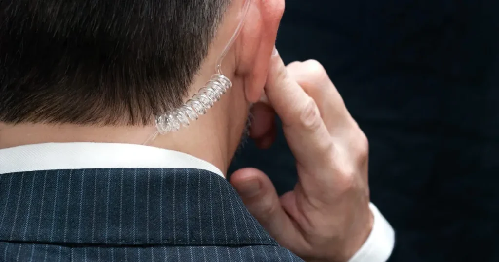 A man in a suit adjusts a clear coiled earpiece, possibly for communication or surveillance.