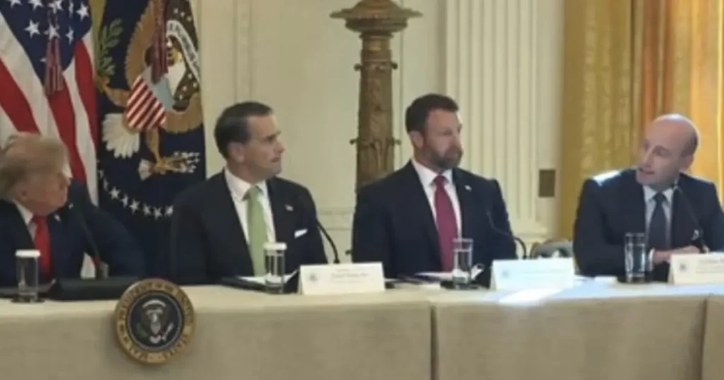 The image shows a formal meeting with four individuals seated at a table, with the U.S. flag and presidential seal in the background.