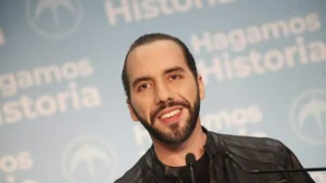The image shows a person speaking at a podium with a backdrop featuring the text "Hagamos Historia."