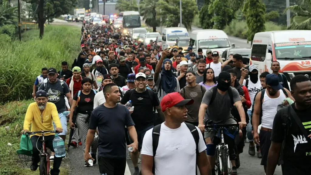 A large group of people, some on bicycles, are walking along a road, possibly participating in a migrant caravan.