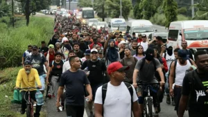 A large group of people, some on bicycles, are walking along a road, possibly participating in a migrant caravan.