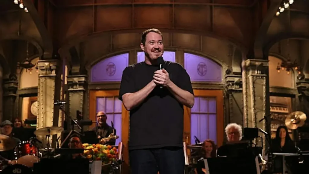 A man stands on stage holding a microphone, smiling, with a band and orchestra in the background.