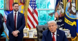 The image depicts a formal setting with two individuals in suits, one seated at a desk and the other standing, in front of a backdrop featuring American flags and military insignia.