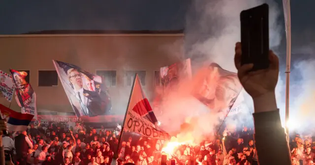 A crowd of people is gathered in front of a building, holding flags and flares, with one person holding up a smartphone to capture the scene.