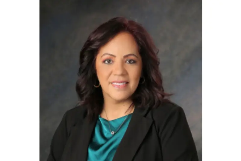 The image shows a professional portrait of a woman with shoulder-length dark hair, wearing a black blazer over a teal top, set against a neutral background.