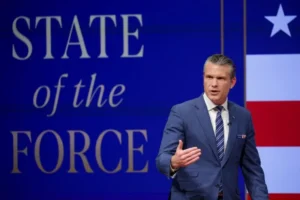 A man in a suit is giving a speech in front of a backdrop that reads "STATE of the FORCE" with an American flag design.