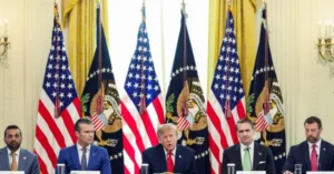 The image shows a formal press conference with several individuals seated at podiums in front of American flags and presidential seals.
