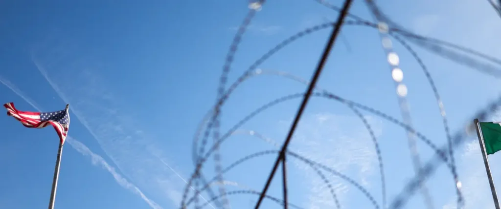 The image shows a barbed wire fence with an American flag on the left and a green flag on the right, set against a clear blue sky.