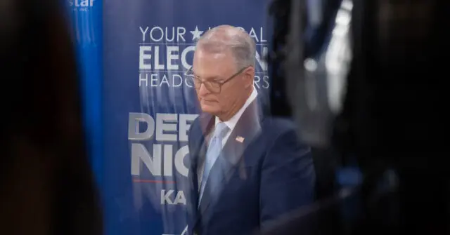 The image shows a man in a suit standing in front of a backdrop with political campaign text.