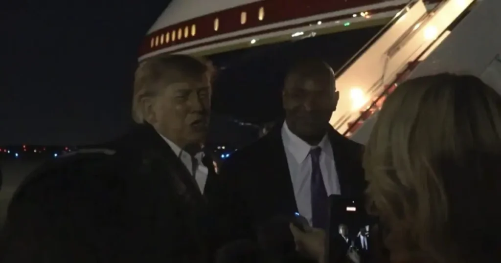 The image shows a man in a suit being interviewed by reporters as he disembarks from an aircraft at night.