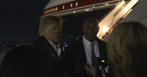 The image shows a man in a suit being interviewed by reporters as he disembarks from an aircraft at night.