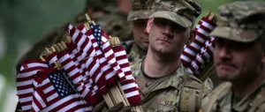 A group of soldiers in camouflage uniforms carry American flags during a solemn ceremony.