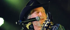 The image shows a musician wearing a cowboy hat and playing a guitar while singing into a microphone on stage.