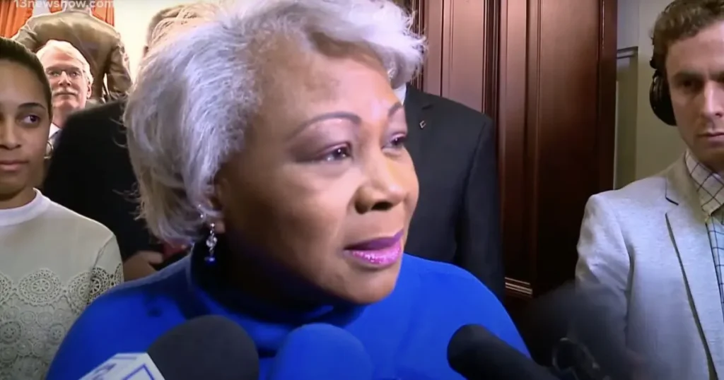 A woman in a blue turtleneck is speaking to reporters surrounded by microphones and other individuals.