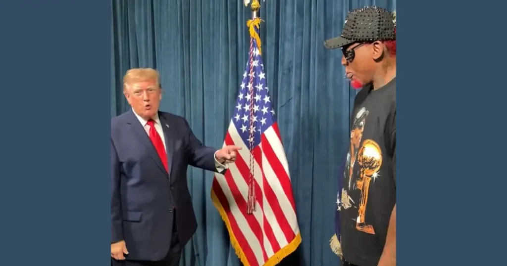 The image shows two individuals standing in front of an American flag, with one person pointing towards the other.