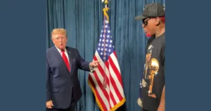 The image shows two individuals standing in front of an American flag, with one person pointing towards the other.