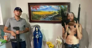 The image shows two individuals holding firearms in a room with a painting of a scenic landscape, a statue of the Virgin Mary, and other religious items.