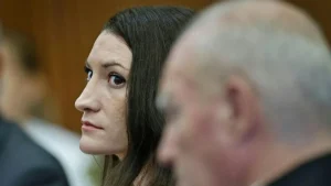 The image shows a woman with dark hair sitting in what appears to be a courtroom setting, with a man in the foreground.