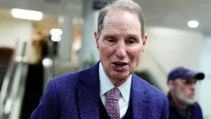 The image shows a man in a suit and tie, speaking in what appears to be a public or professional setting.
